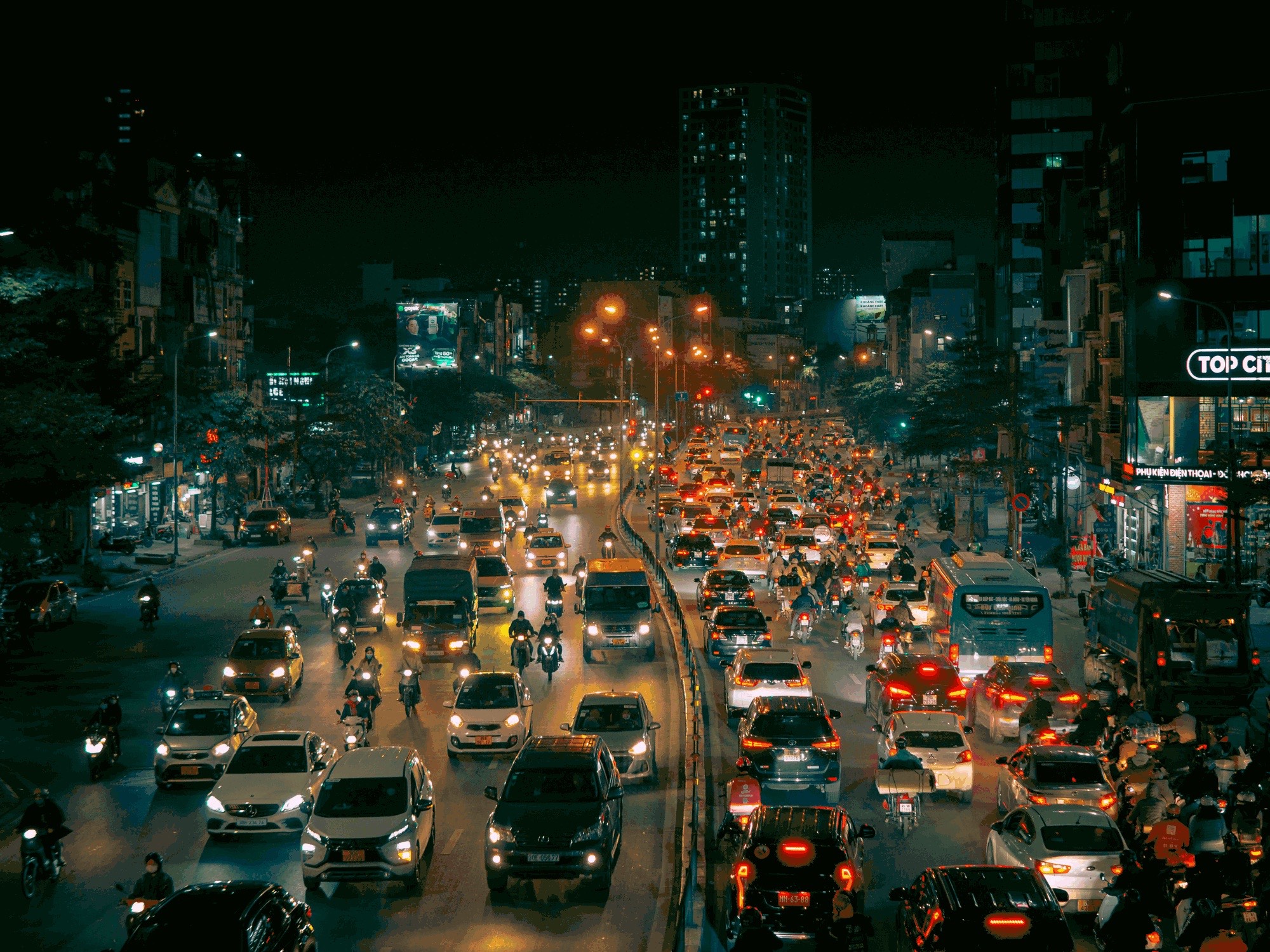 A congested city road at night seen from above: dense lanes of cars, buses and motorbikes crawl between lit shopfronts and high-rises.