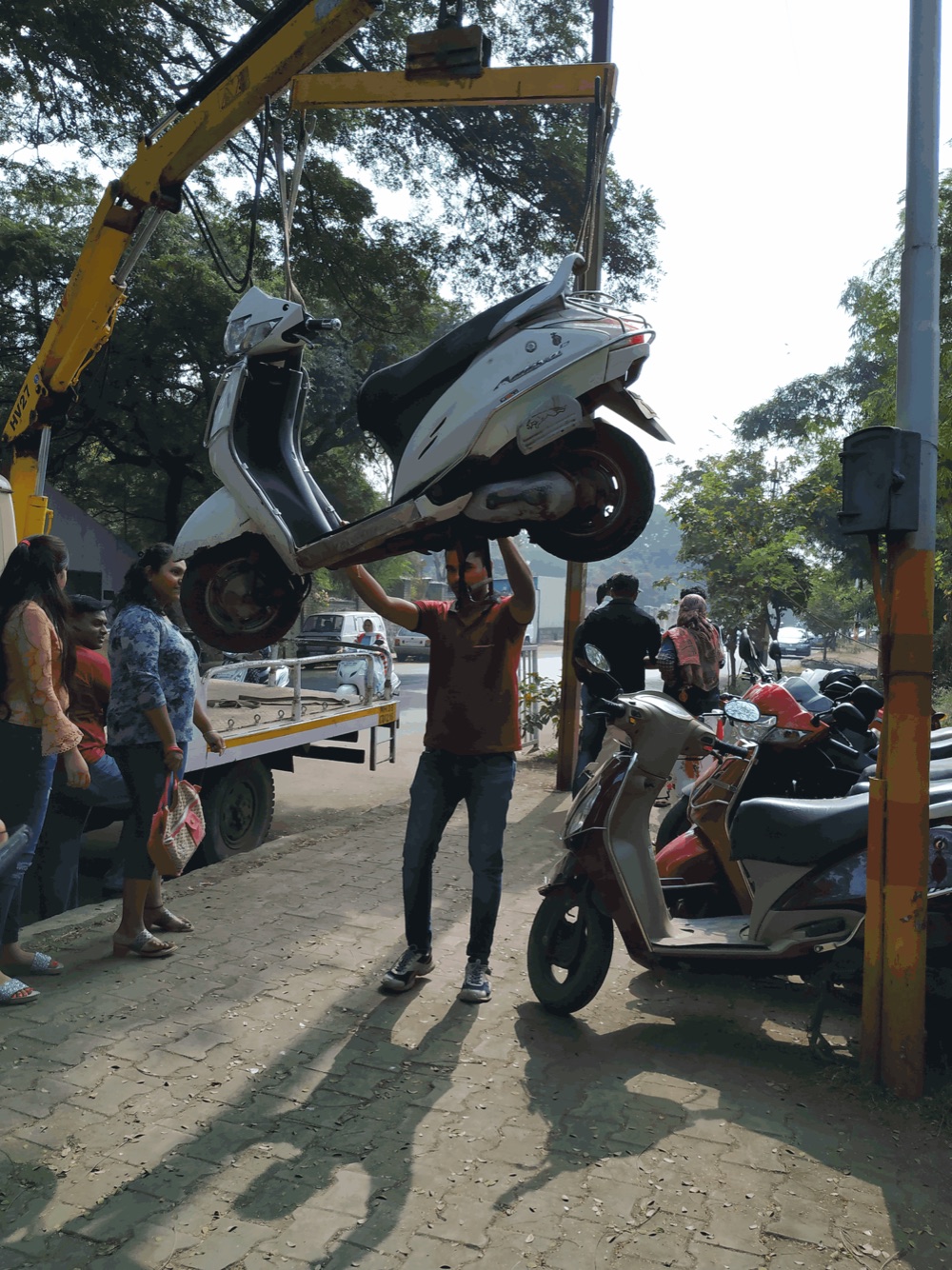 A tow crane lifts a white scooter off the pavement as a man reaches up beneath it and onlookers stand beside the tow truck.